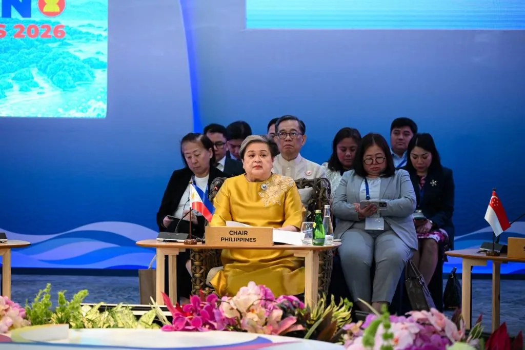 A woman in a gold embroidered dress sits at a 'CHAIR PHILIPPINES' desk during a formal 2026 summit, with the Philippine flag and officials visible behind pink orchids.