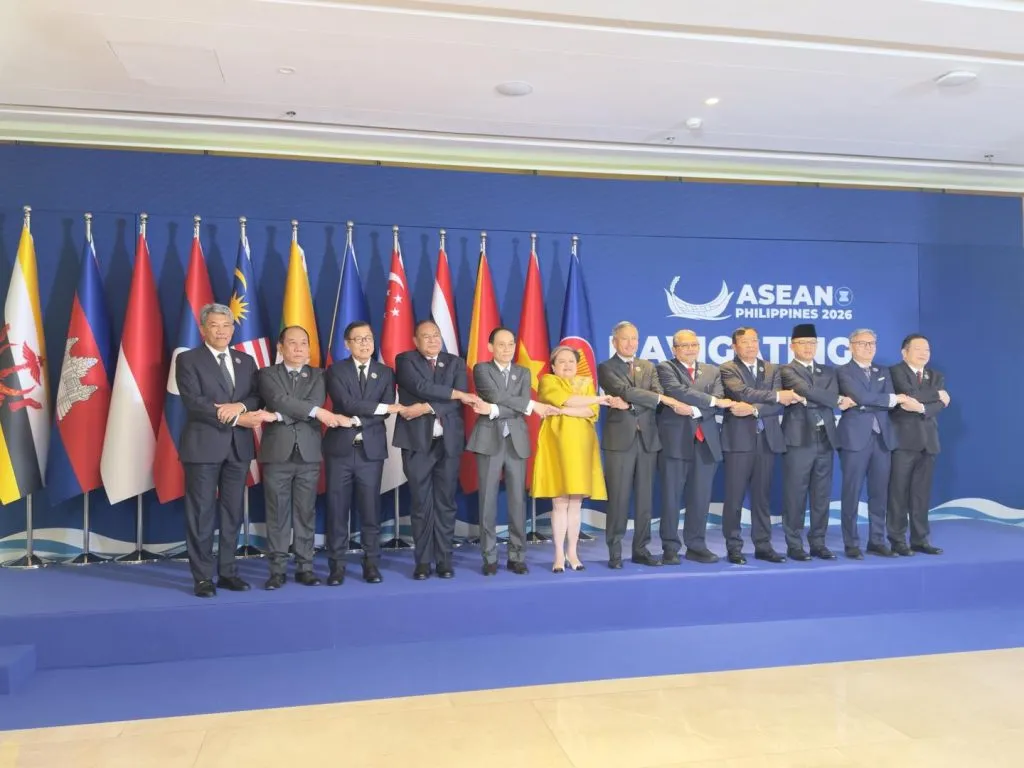 Diplomats in formal suits stand in a long row on a blue platform, performing a traditional cross-arm handshake. Colorful national flags and a formal logo decorate the backdrop.