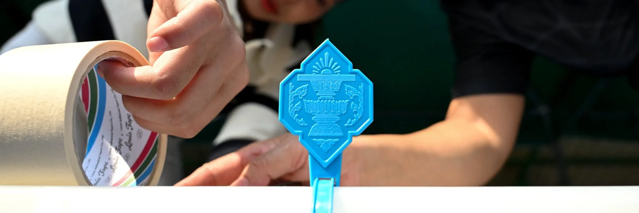Two people carefully seal a white container using a roll of masking tape. A bright blue plastic security clip with a detailed emblem secures the lid's edge.