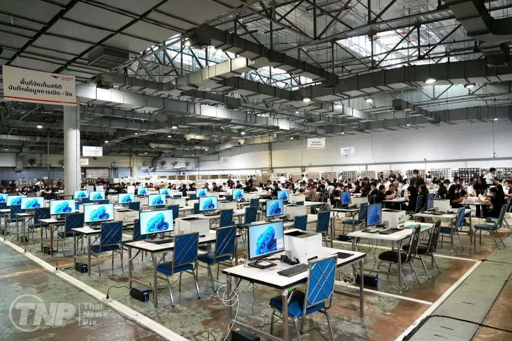 A cavernous, industrial hall filled with rows of computer stations and blue chairs. In the distance, dozens of workers sit at long tables, meticulously sorting through large stacks of paperwork.