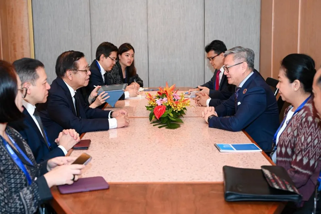 Professionals in formal business attire sit across from each other at a long conference table, engaged in a serious meeting. A bright floral centerpiece sits between the two groups.