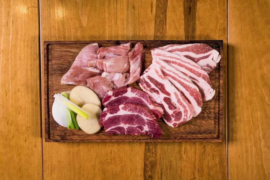 On a warm wooden table, a cutting board holds raw pork belly, marbled shoulder cuts, potatoes, and green onions. A fresh meal awaits preparation.