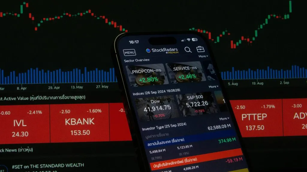 A smartphone displaying a financial app sits before a computer screen filled with glowing red and green candlestick charts and market indices, creating a dense, data-driven scene.