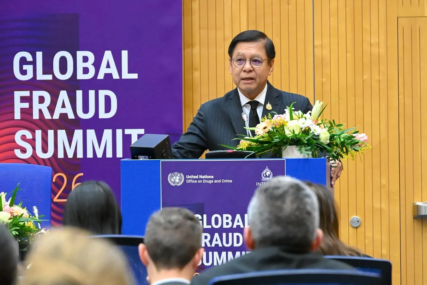 A speaker in a dark pinstripe suit delivers remarks at the Global Fraud Summit from a blue podium with flowers before a formal audience.