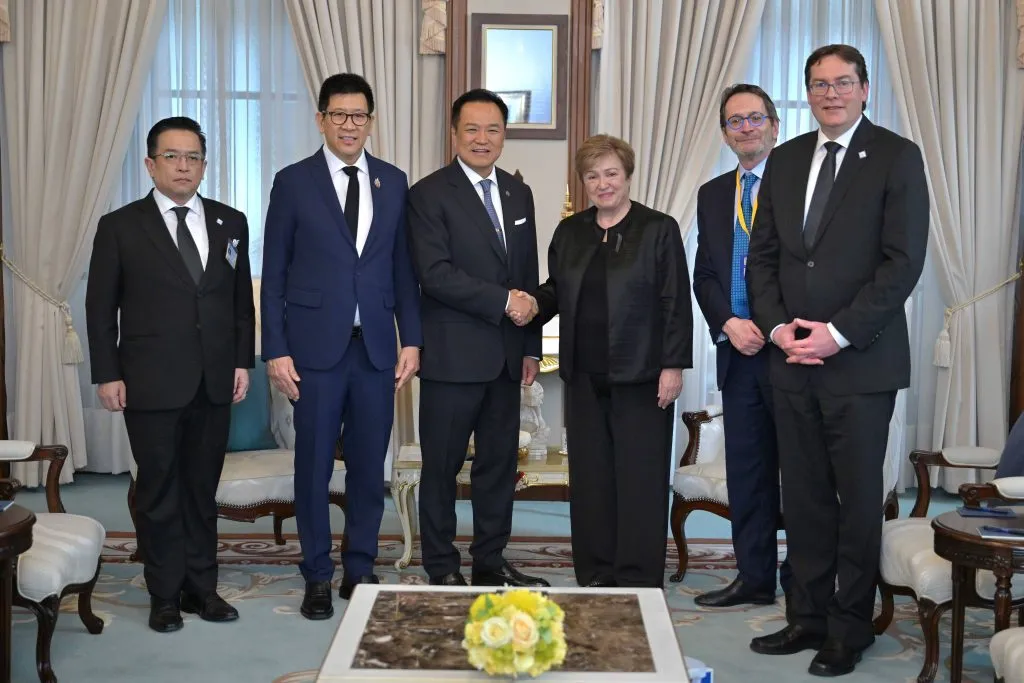 Six officials in formal attire stand in an elegant room. Centrally, a Thai man shakes hands with a woman. Ornate curtains and chairs frame this important diplomatic meeting.