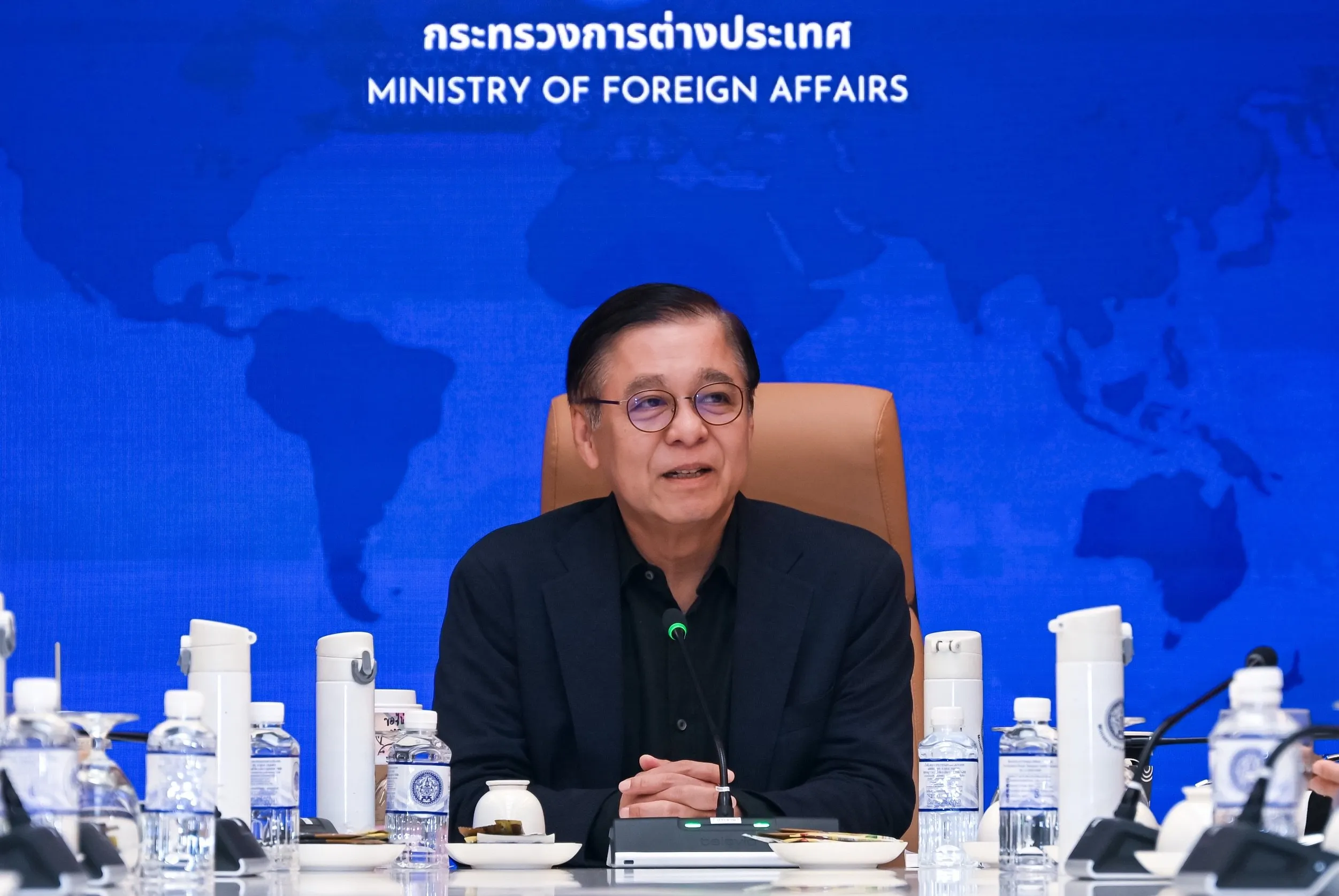 A man wearing glasses sits at a table with water bottles. Behind him is a blue world map backdrop with 'Ministry of Foreign Affairs' displayed in white text.