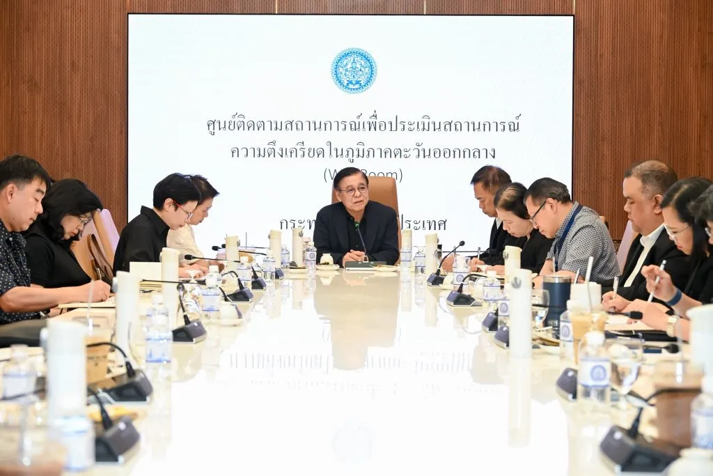 A formal meeting in a boardroom. Several people sit at a long white table with microphones, while a man at the head of the table leads the session.