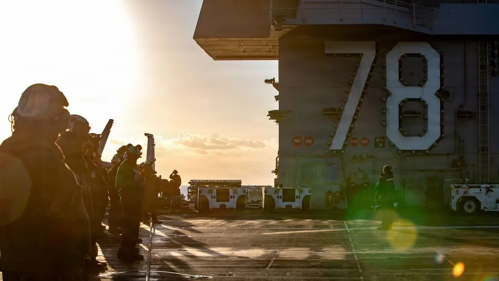 Officers gather on a flight deck at sunset, silhouetted against the glowing sky. The massive aircraft carrier island, labeled "78," overlooks military personnel and equipment at sea.