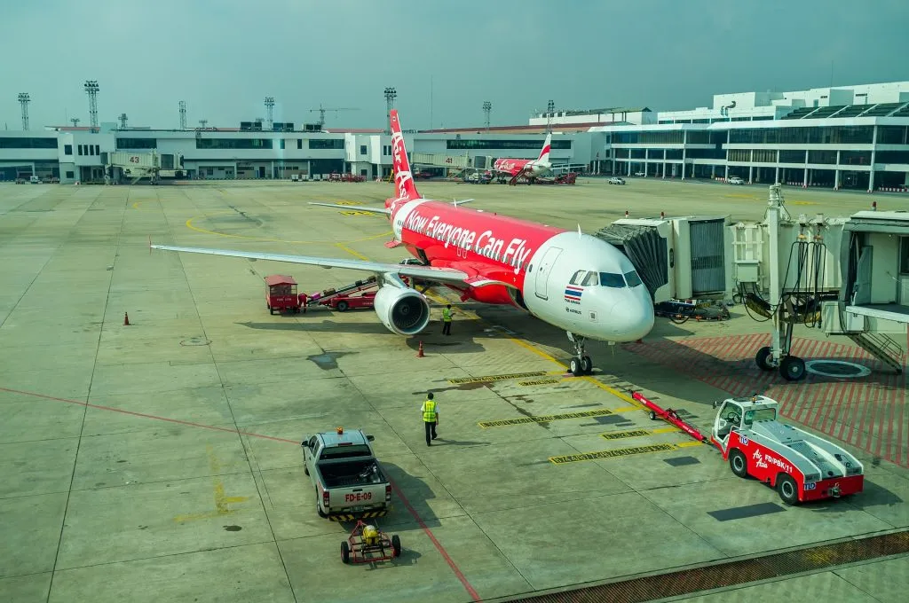 A red Thai AirAsia airplane sits at a jet bridge on a bright airport tarmac. Ground crew members in yellow vests walk near various vehicles and terminal buildings.