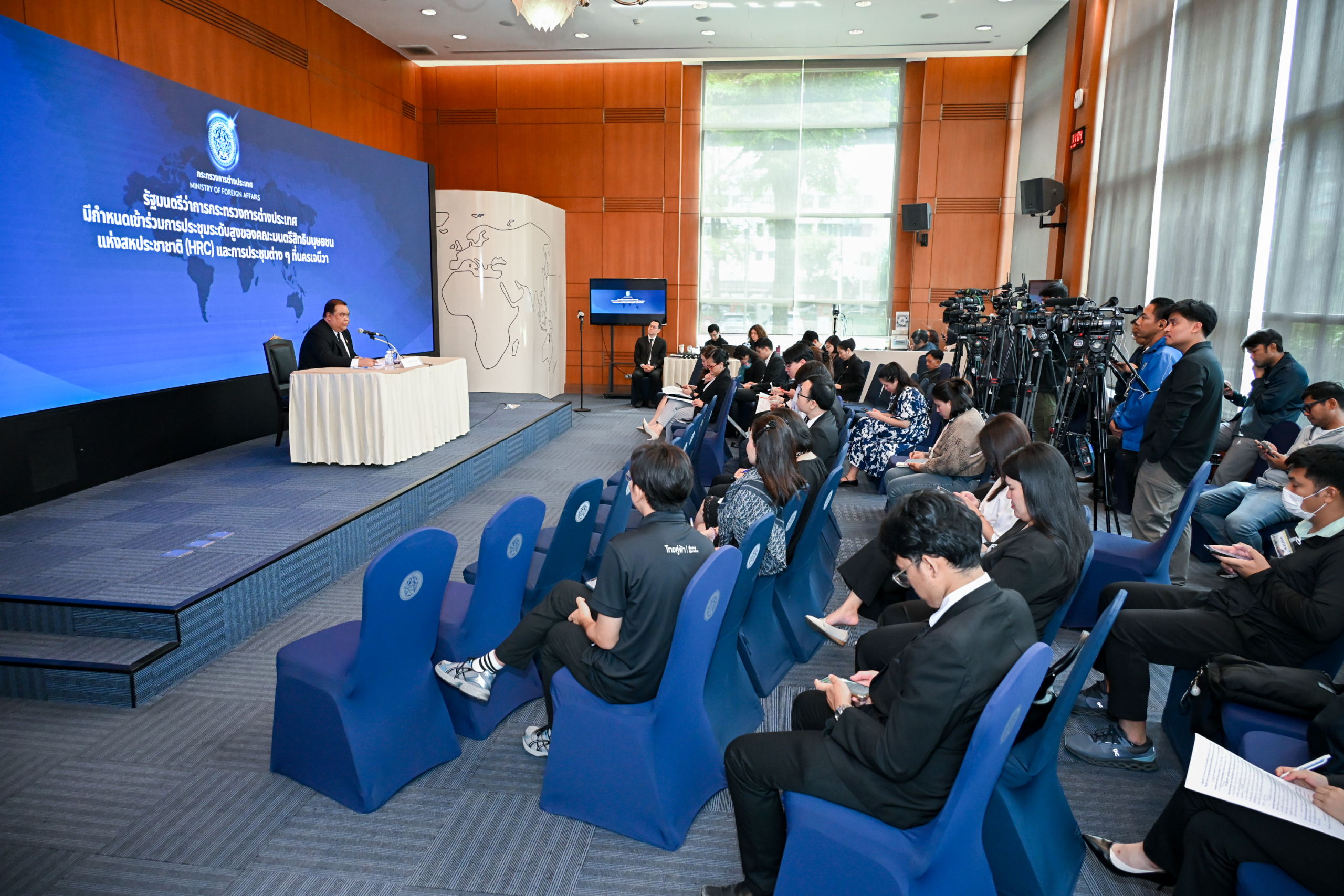 A speaker sitting on a stage in front of a school of reporters.