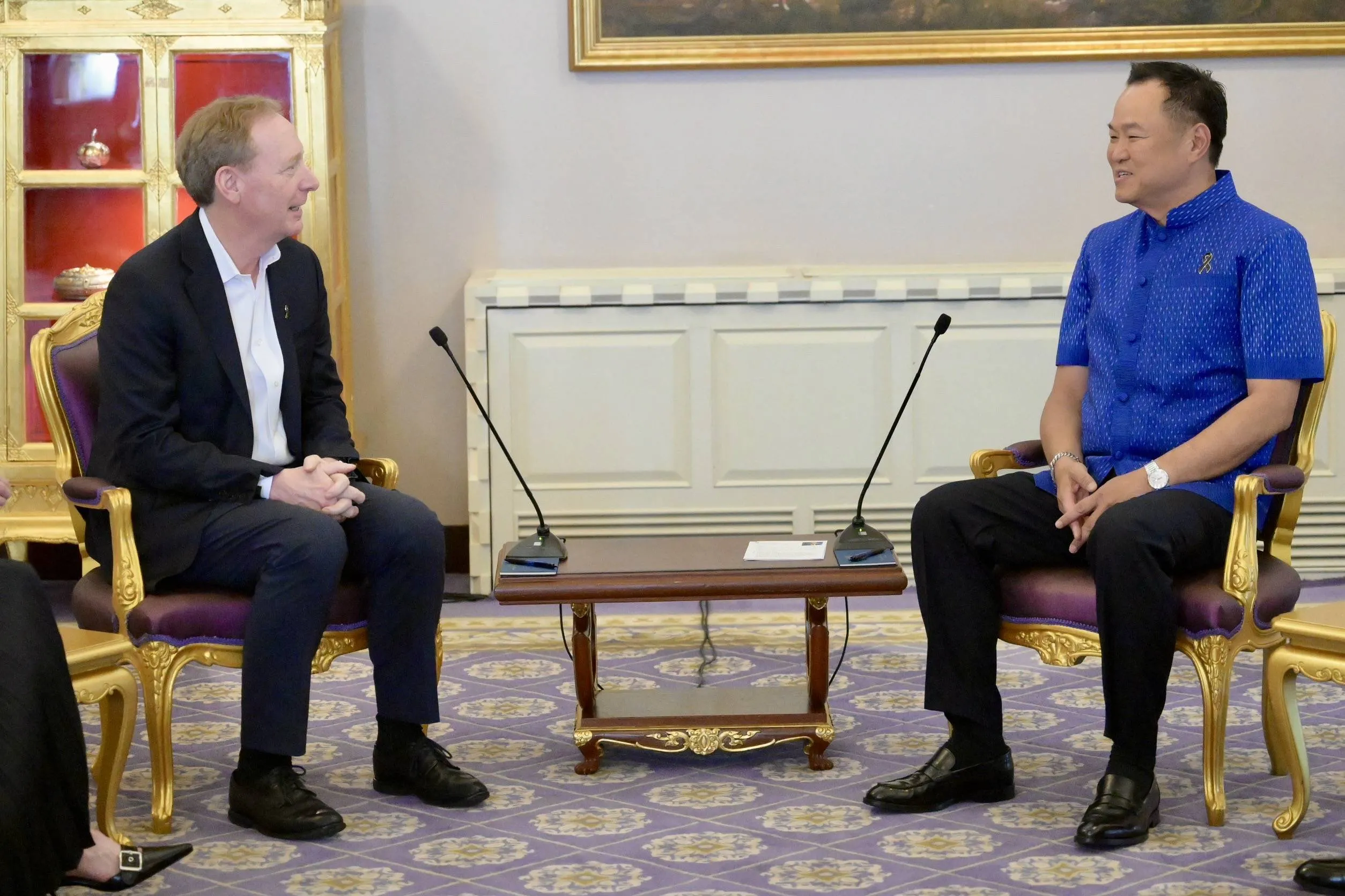 In a formal room, two men sit in ornate gold chairs. One wears a suit; the other, a vibrant blue traditional Thai shirt.