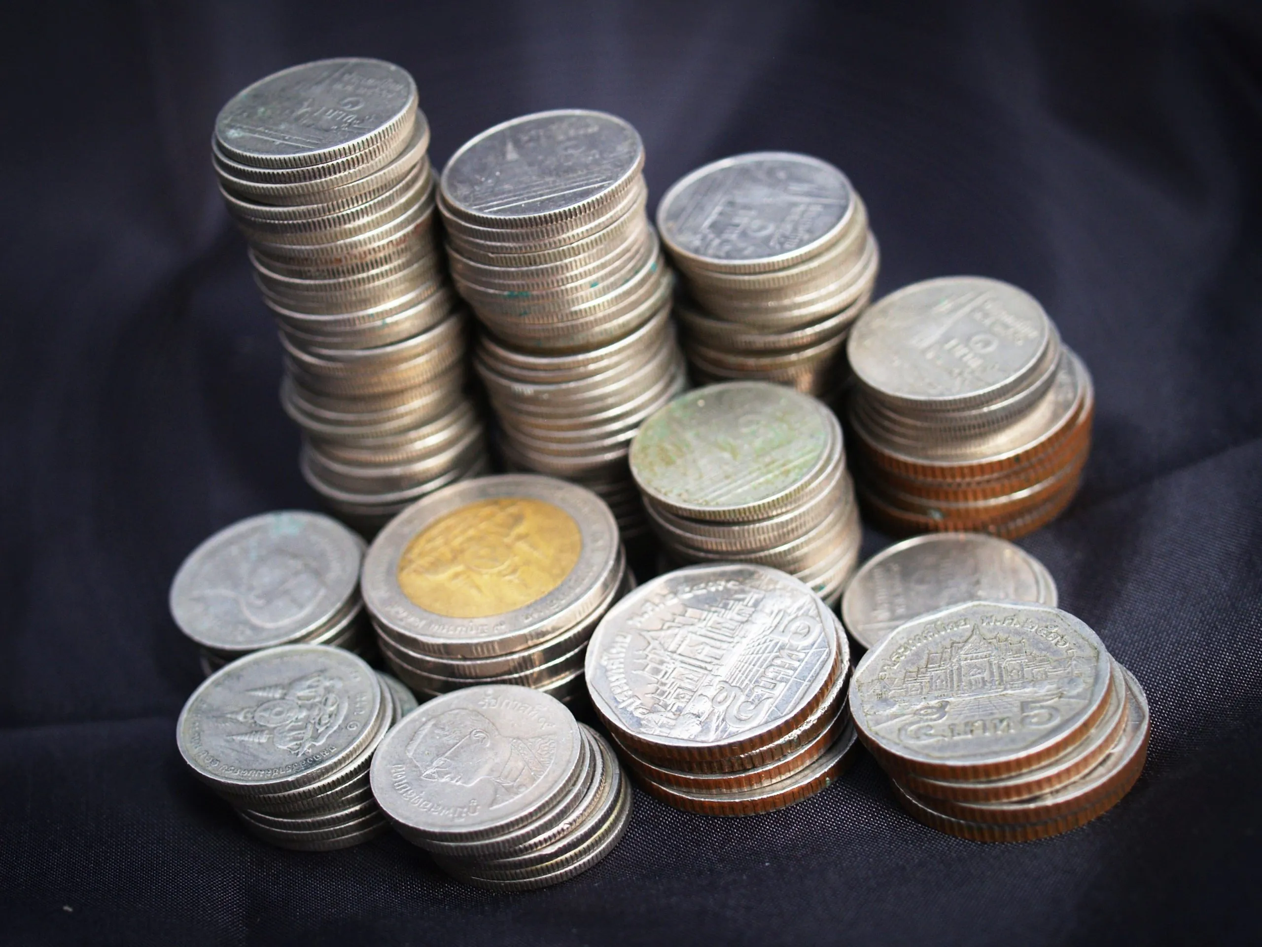 Various Thai baht coins—silver, bronze, and gold-rimmed—stacked in small piles and scattered across a dark fabric surface, embossed with temple designs and numerals.