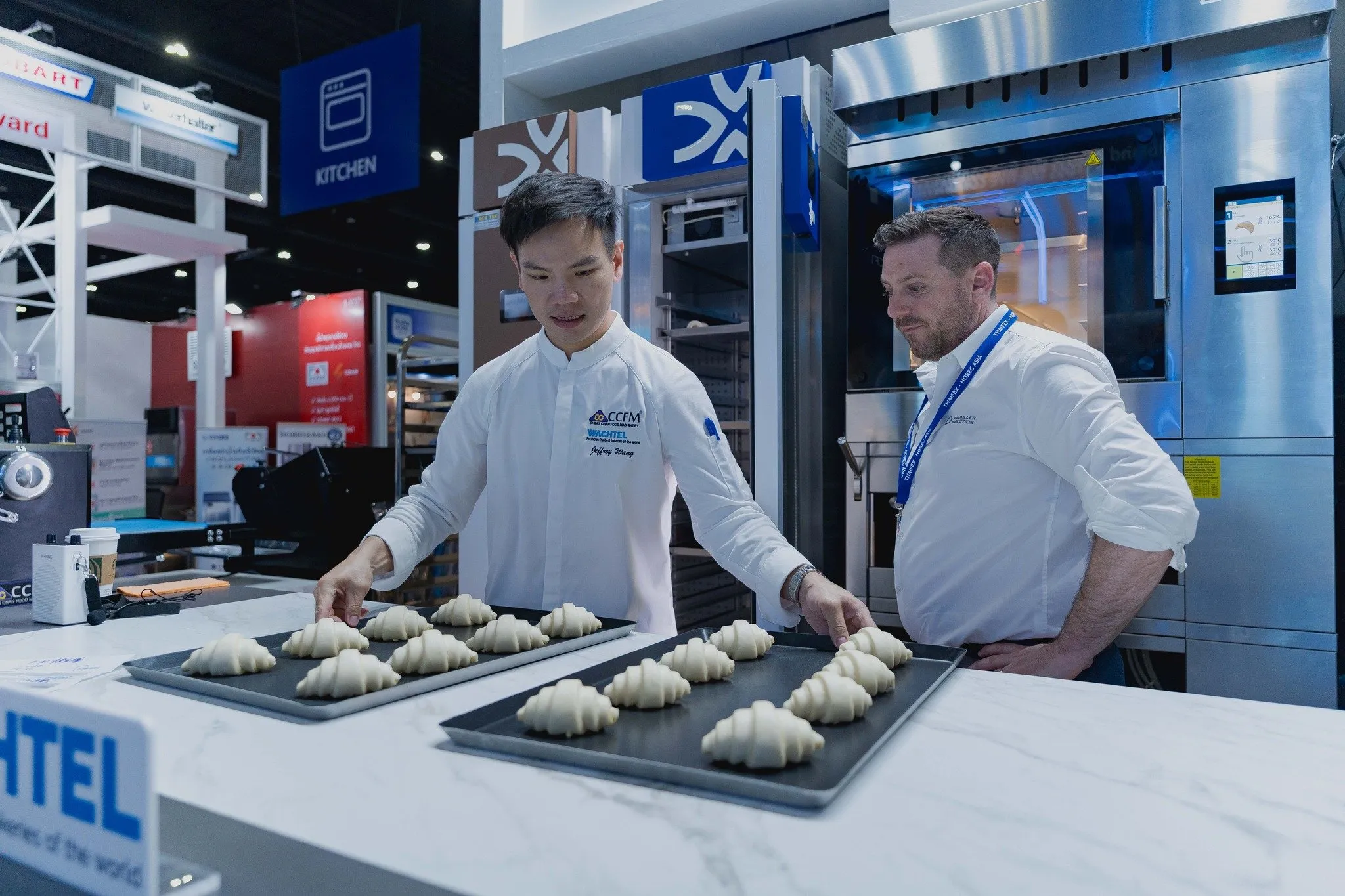 A chef in white coat arranges unbaked croissants on a marble table while a man observes, beside a stainless steel oven under blue KITCHEN signage.