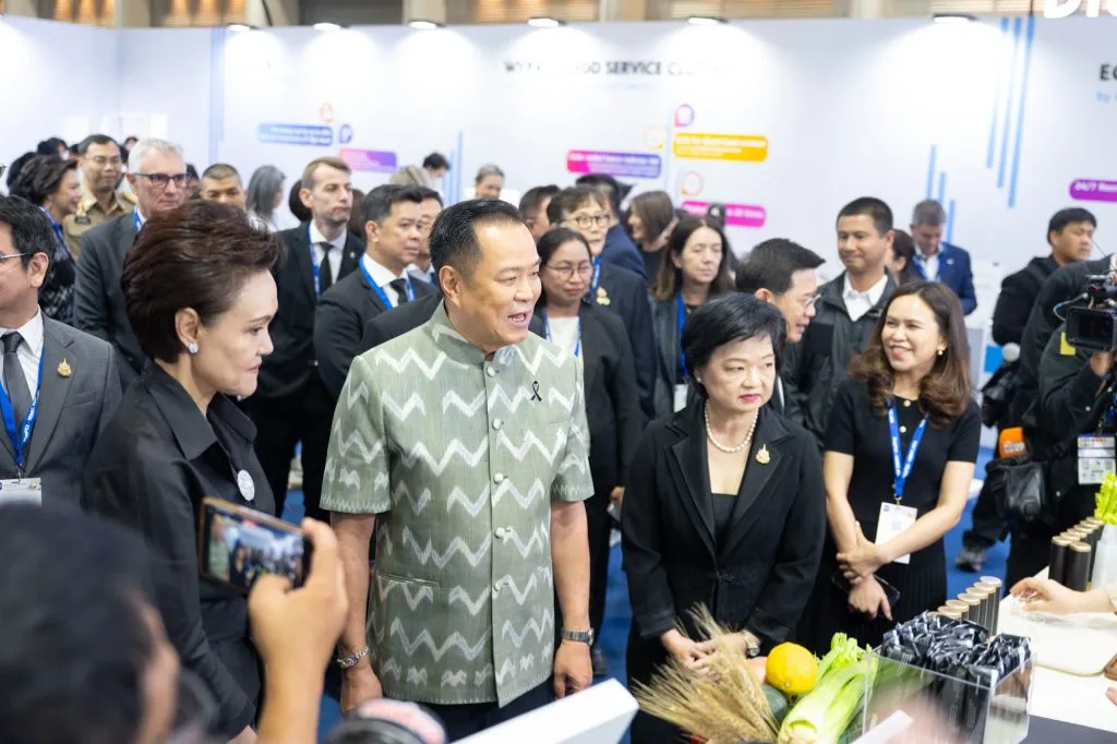 A man in a green chevron-patterned shirt talks at a crowded trade show booth, flanked by officials in formal black suits and spectators.