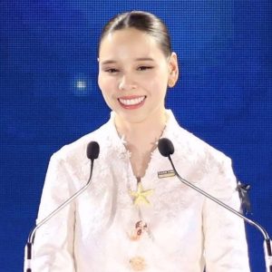 A woman in a white blouse with a starfish brooch smiles behind a "Thailand Biennale Phuket 2025" podium against a vibrant blue backdrop.