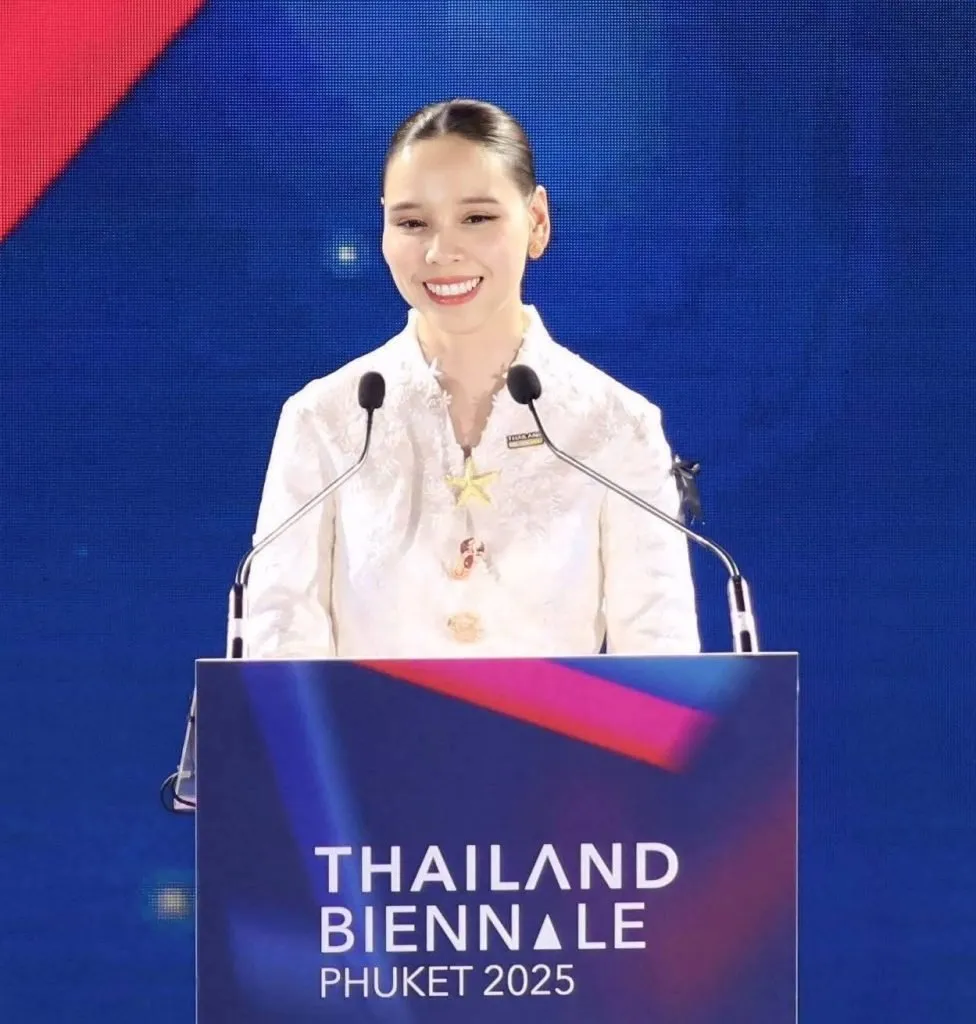 A woman in a white blouse with a starfish brooch smiles behind a "Thailand Biennale Phuket 2025" podium against a vibrant blue backdrop.
