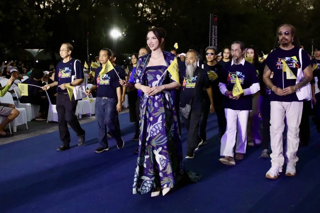 A woman in a patterned blue gown leads a procession on a blue carpet, flanked by people in event T-shirts waving small yellow flags.