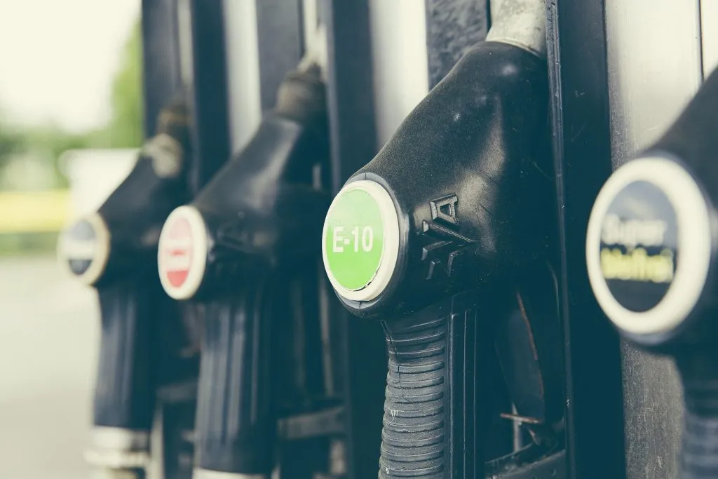 Several black fuel pump handles hang in a row. The closest one features a prominent green circular label with white text 'E-10' against a blurred background.