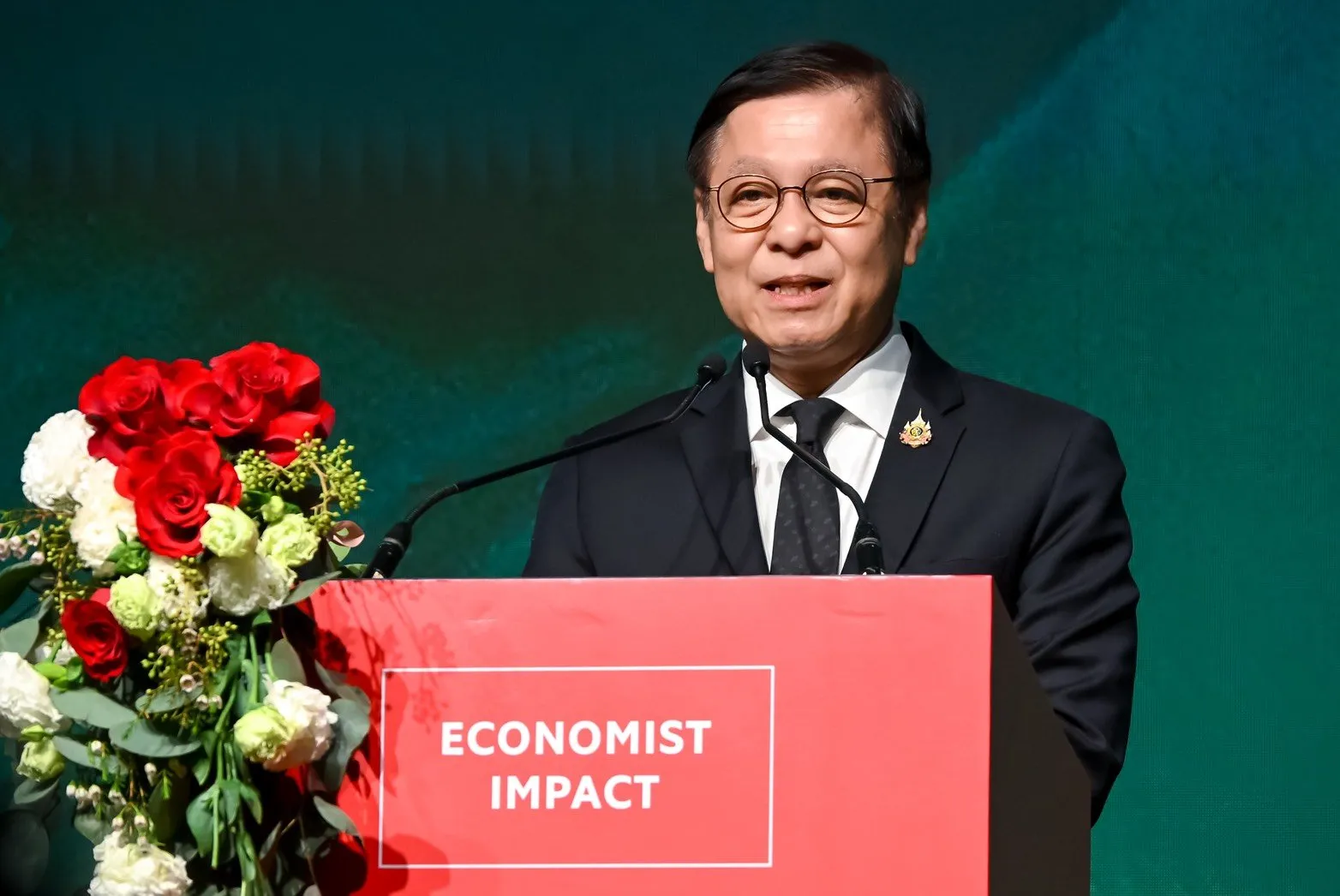 A smiling Asian man with glasses speaks from a red "Economist Impact" podium, flanked by red roses against a dark green background.