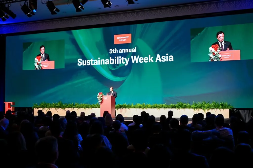 A man speaks at the "5th annual Sustainability Week Asia." A massive screen towers over him, while a large audience sits in silhouetted rows.