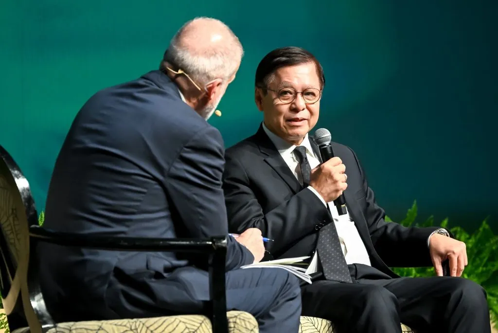 Seated on stage, a man in a suit speaks into a handheld microphone toward an interviewer, both positioned before a solid green background.
