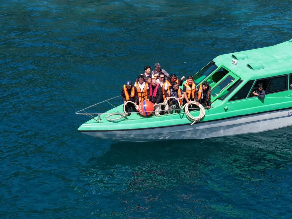 A group of men in orange life jackets stand on the deck of a green boat, holding ropes and a red buoy amidst deep blue water.