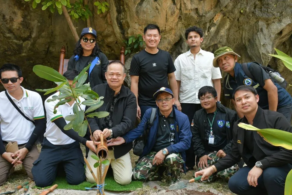 Ten people are gathered outdoors for a tree planting event near a massive rocky cliff. Five men kneel around a sapling while others stand behind them.