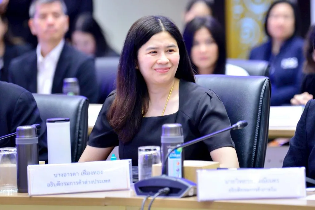 A woman with long black hair sits at a conference table, smiling warmly. She wears a black dress and stands behind a formal nameplate.