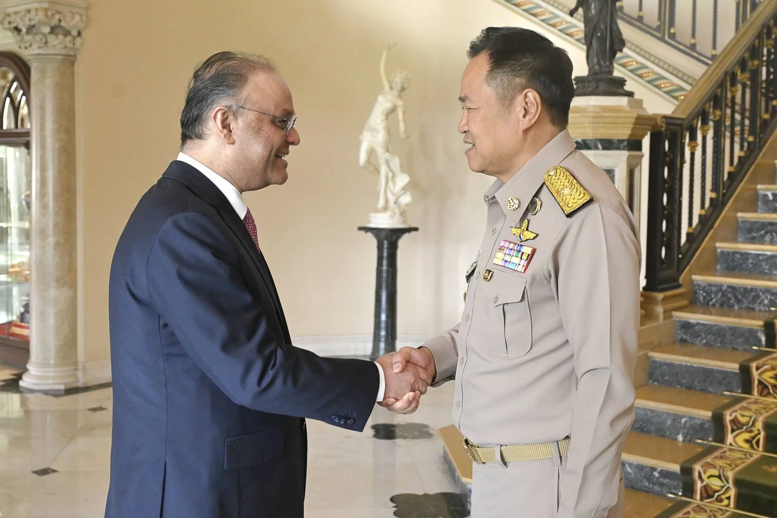 Two men, one in a suit and one in khaki uniform, shake hands before a grand, ornate double staircase in an elegant, formal hall.