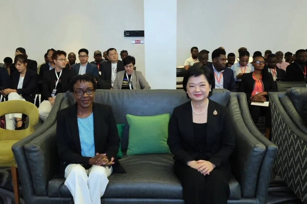 An Asian woman in black and a Black woman in stripes sit on a grey sofa, facing forward before a seated audience.