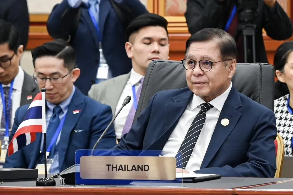 A man in a suit and tie sits behind a "THAILAND" nameplate at a formal conference, with colleagues and a small Thai flag nearby.