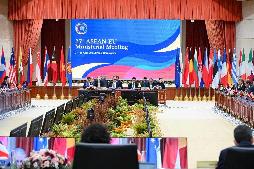 A large conference hall features officials seated at long tables, flanked by numerous international flags under a digital screen for the 25th ASEAN-EU Ministerial Meeting.