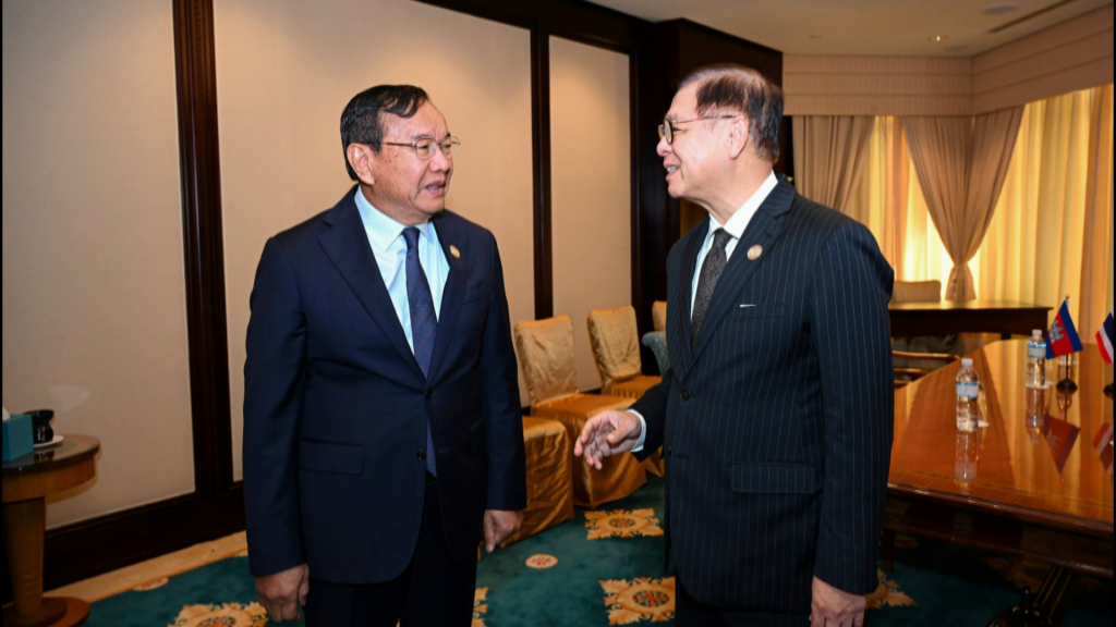 Two men in professional suits and glasses stand in a meeting room, engaged in conversation near a wooden table with small national flags.