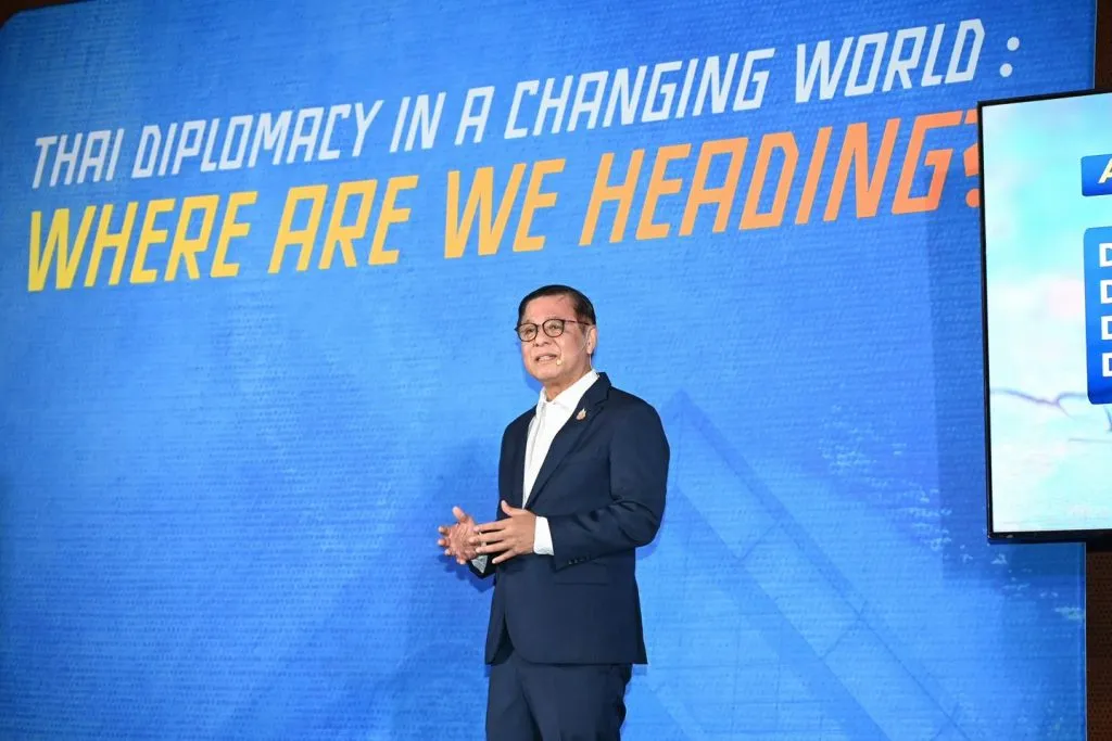 A man in a navy suit speaks on a blue stage before a large banner reading: "THAI DIPLOMACY IN A CHANGING WORLD: WHERE ARE WE HEADING?"