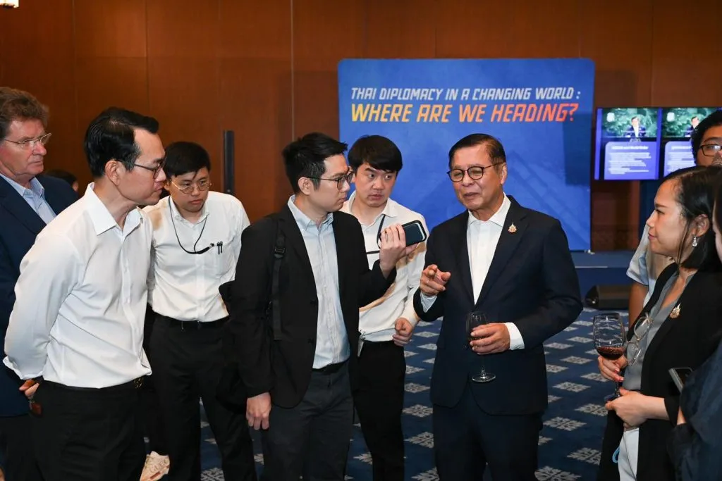 A man in a suit addresses a group, holding a wine glass. One man records him with a phone. The diplomacy banner remains behind them.