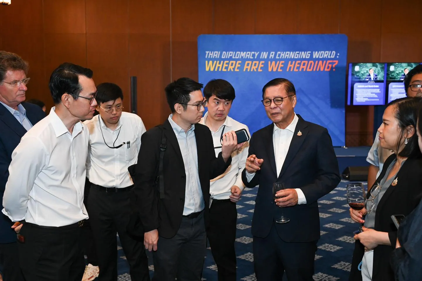 A man in a suit addresses a group, holding a wine glass. One man records him with a phone. The diplomacy banner remains behind them.