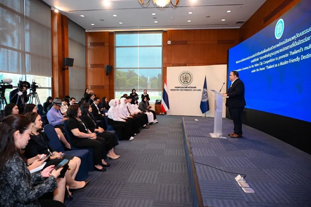 A man speaks at a podium in a formal hall before seated guests and a blue screen promoting Thailand as a "Muslim-Friendly Destination."