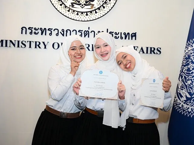 Three smiling young women in white hijabs and uniforms pose with award certificates in front of a Ministry of Foreign Affairs sign in Thailand.