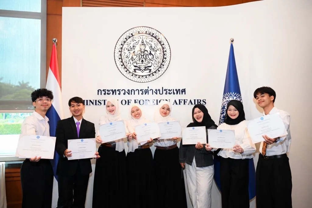 Eight smiling young adults pose with award certificates in front of a Thai Ministry of Foreign Affairs sign, flanked by national and ministry flags.