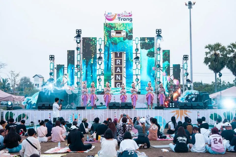 Seven traditional Thai dancers perform on a vibrant, illuminated outdoor stage under a "Smile Trang" sign. An audience sits on the grass watching the performance.