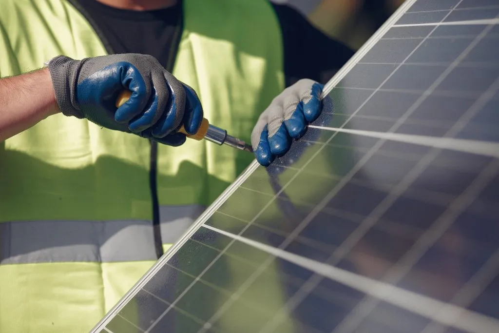 A worker in a neon safety vest and blue gloves uses a tool on a solar panel’s frame under clear, direct sunlight.