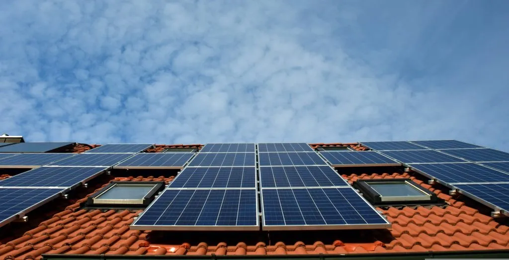 Blue solar panels cover a terracotta tiled roof with skylights, positioned under a blue sky filled with a pattern of small, puffy white clouds.