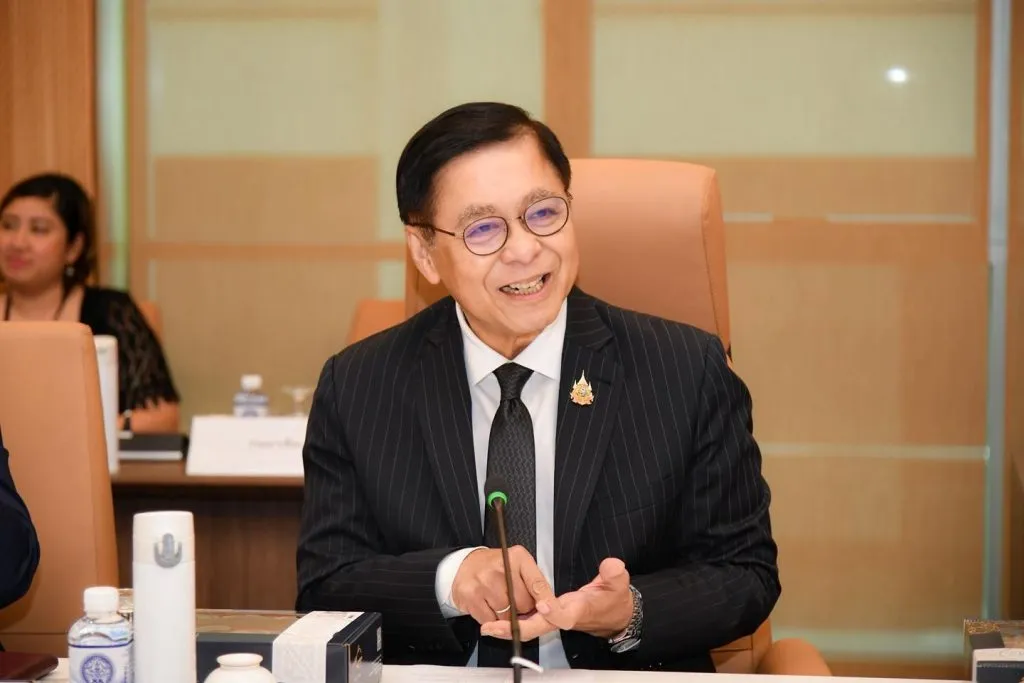 A smiling man in a pinstripe suit and glasses gestures during a meeting, seated in a professional, tan-colored chair.
