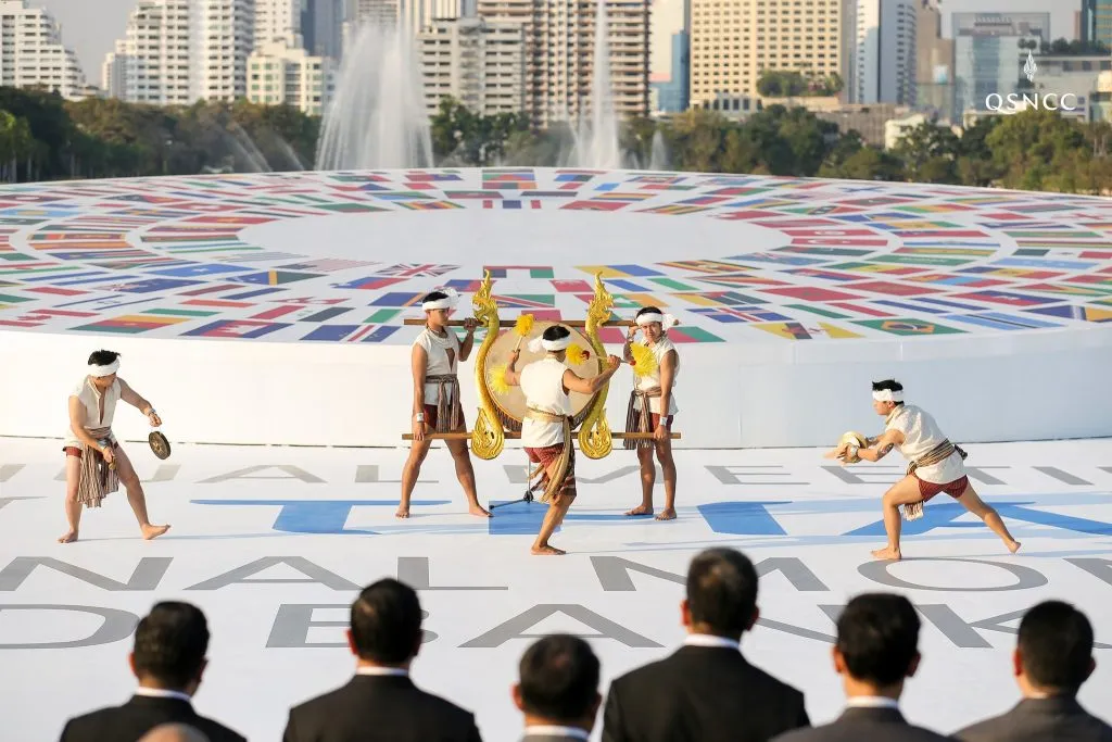 Thai performers in traditional attire play drums on a white stage decorated with global flags and event text.