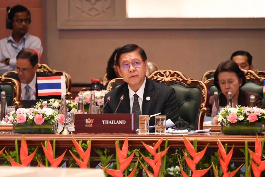 A middle-aged Thai man in a dark pinstripe suit and glasses sits at a conference table behind a "THAILAND" nameplate, speaking intently.