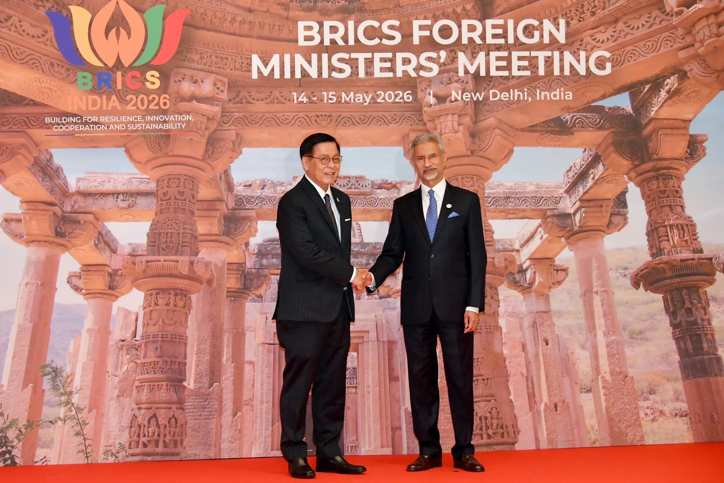 A Thai official shakes hands with an Indian diplomat in front of a large backdrop featuring ancient Indian temple pillars.