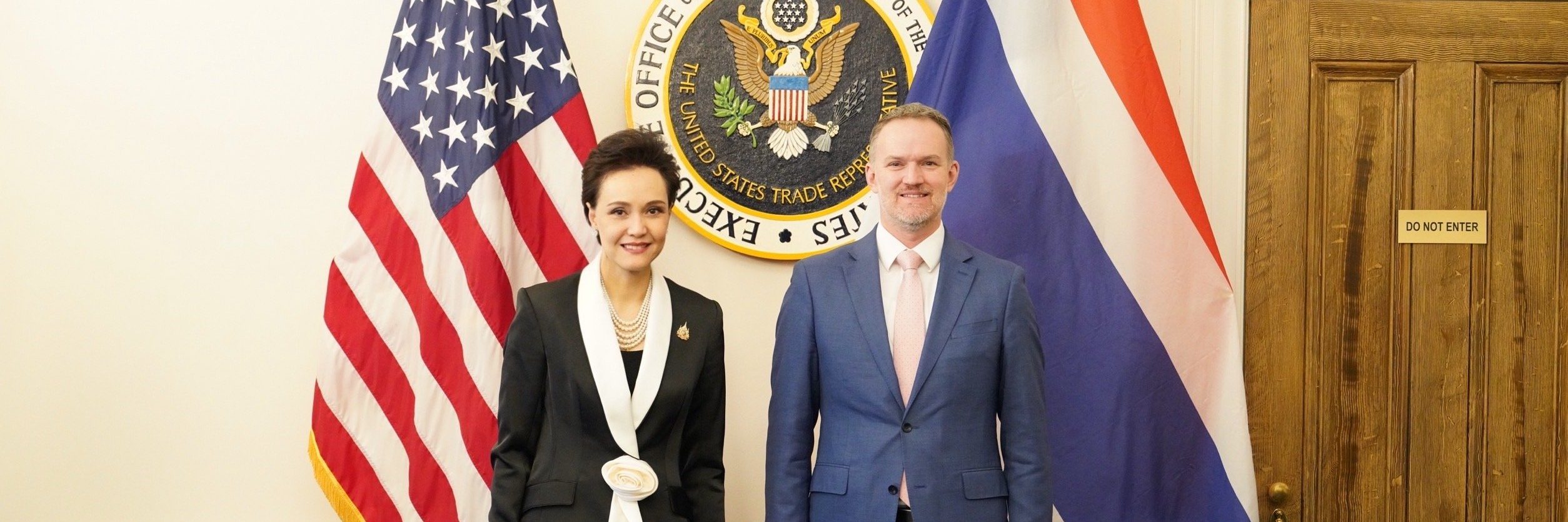 A Thai woman in a black suit with a white sash stands beside a male US official between the American and Thai flags.