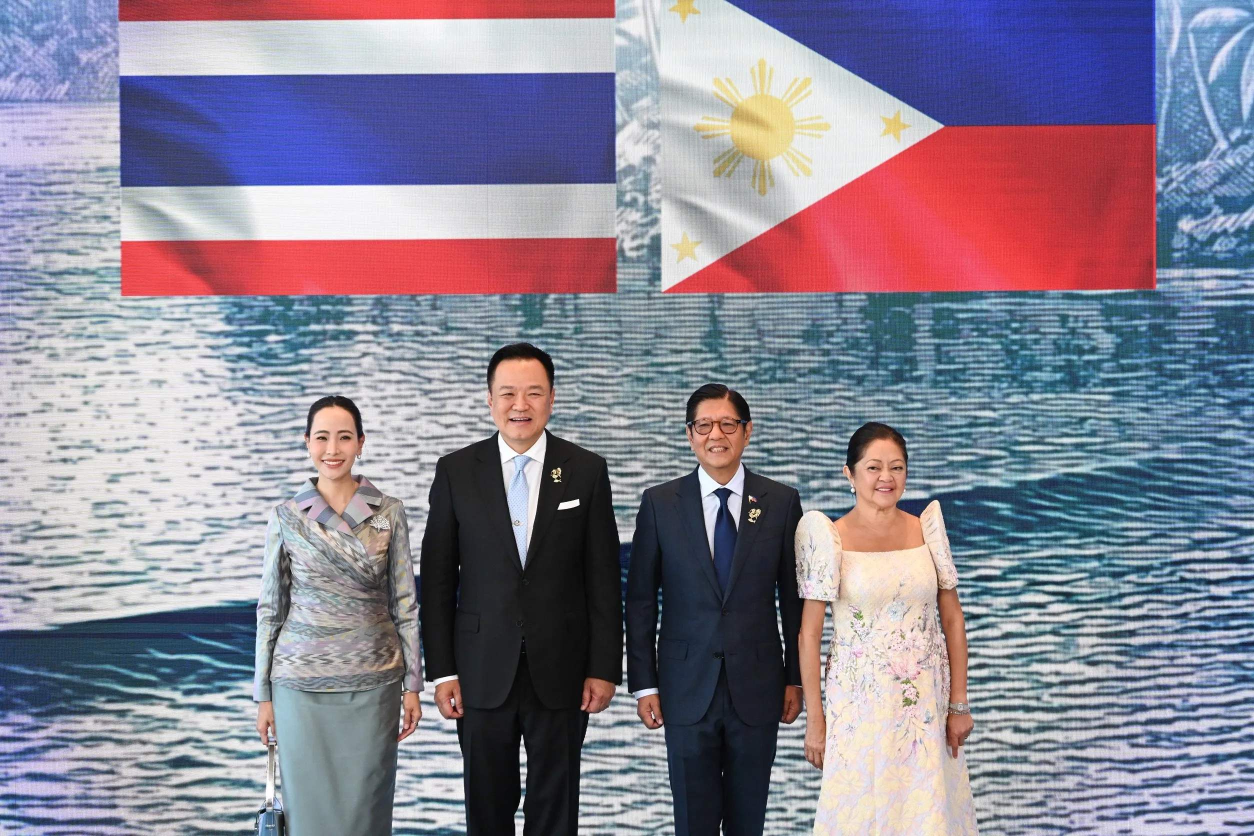 Two couples stand before flags of Thailand and Philippines; men in dark suits, women in elegant traditional floor-length dresses.