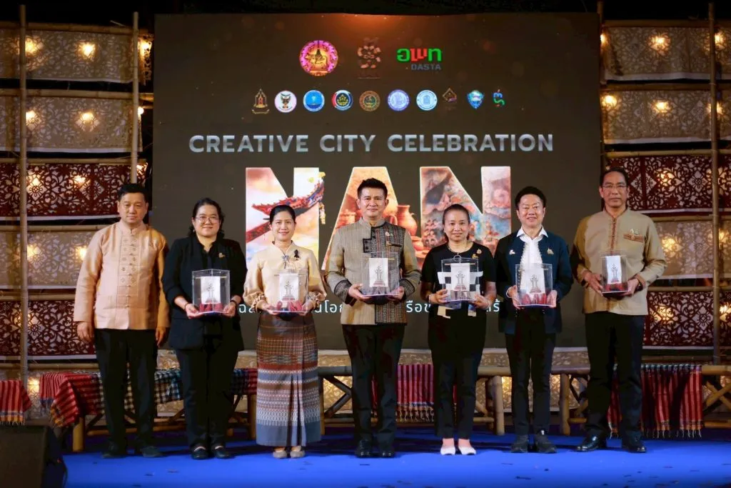 Seven officials stand onstage holding crystal awards before a large screen reading "CREATIVE CITY CELEBRATION NAN," flanked by woven bamboo panels with warm lighting.