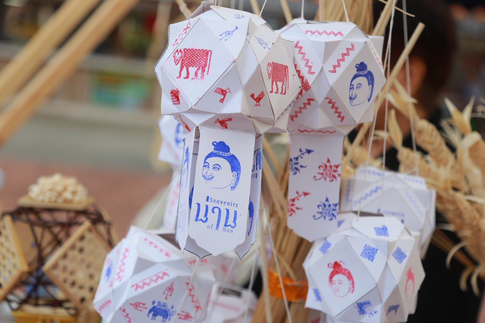 Close-up of geometric white paper lanterns decorated with blue and red stamps of traditional figures, animals, and Thai text reading "Souvenirs of Nan."